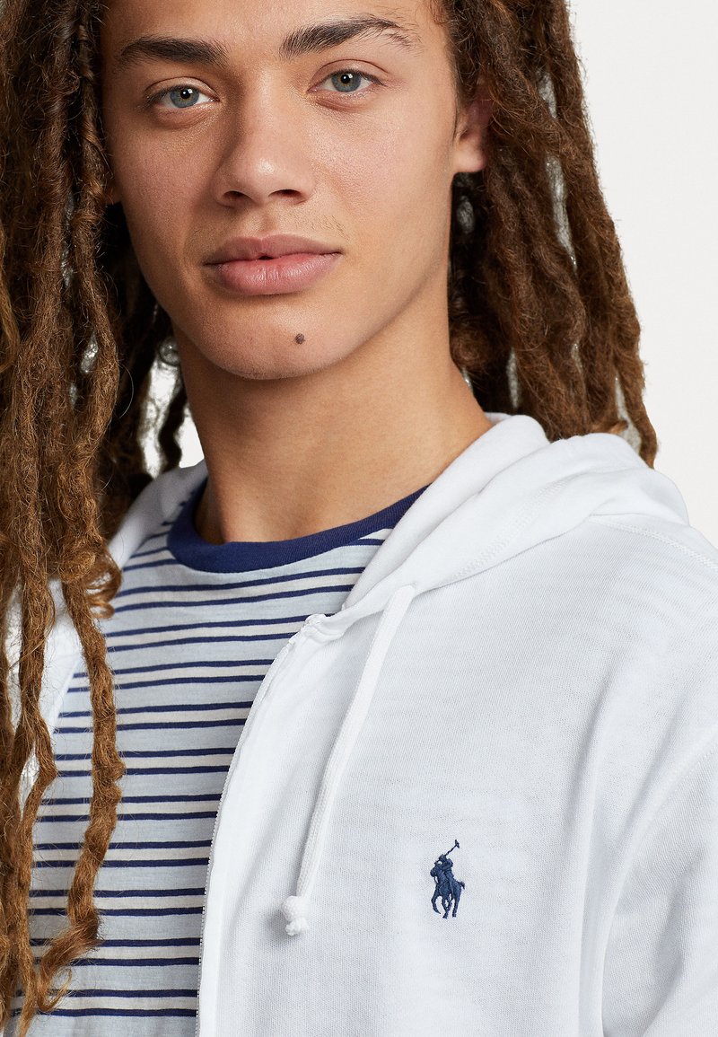Weiße Zip-Hoodie mit Kordelzug, ausgestattet mit einem kleinen dunkelblauen Logo. Darunter ist ein grau- und dunkelblau gestreiftes T-Shirt sichtbar.