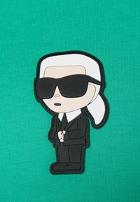 Sørøver gummi-patch af en figur i en sort jakkesæt med en hvid skjorte og solbriller, med hvidt hår, på en grøn tekstureret stofbaggrund.