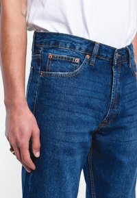 Jeans de mezclilla en un lavado azul oscuro con un corte recto, que presentan remaches de cobre, costuras naranjas y bolsillos laterales.