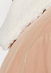 Beige ytterplagg med en texturerad vit fleecekrage. Slät tyg med synliga sömnadsdetaljer på sidorna. Mjuk, varm utseende.