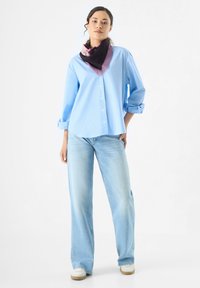Chemise bleu clair à manches roulées, associée à un jean bleu clair et à un foulard coloré aux teintes violettes, noires et roses.
