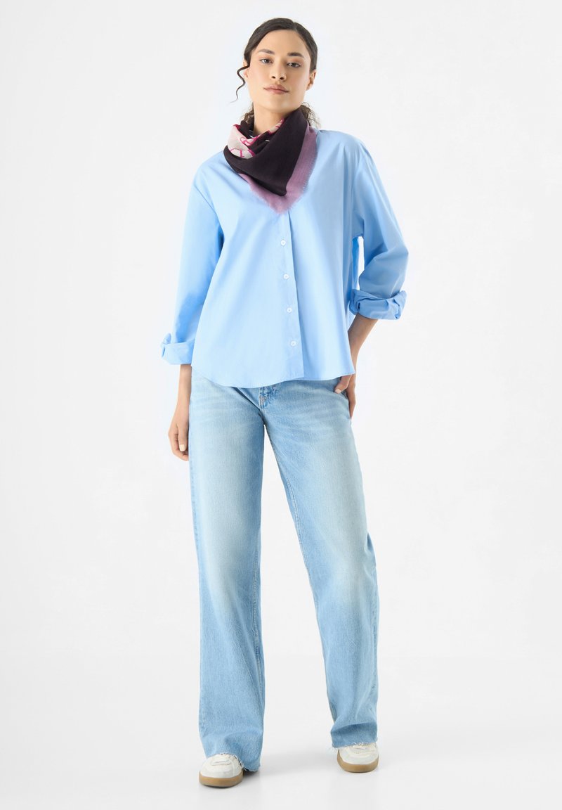 Chemise bleu clair à manches roulées, associée à un jean bleu clair et à un foulard coloré aux teintes violettes, noires et roses.