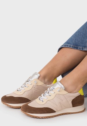 Persoon draagt beige en bruine sneakers met witte veters en zilveren en gele accenten, gecombineerd met blauwe spijkerbroek.