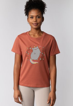 Jeune femme portant un t-shirt couleur rouille avec un chat licorne dessin animé entouré d'éléments floraux, debout devant un fond gris clair.