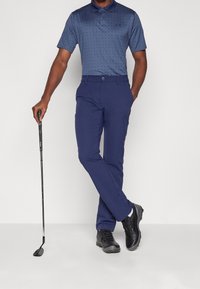 Polo bleu marine avec motif à pois, associé à un pantalon bleu marine. Le modèle tient un club de golf et porte des chaussures noires, affichant une tenue sportive.