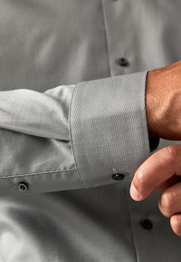 Grau strukturiertes Hemd mit Button-down-Kragen, schwarzen Knöpfen und einer Nahaufnahme des Ärmels mit glattem Finish und dezentem Muster.