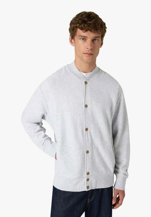 Giovane uomo con capelli ricci che indossa una felpa grigia chiara con bottoni, in piedi con una mano in tasca e jeans scuri.