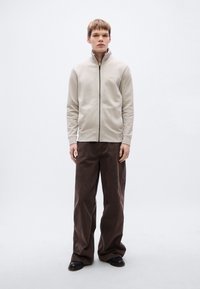 Φωτεινό μπεζ φούτερ με φερμουάρ και όρθιο γιακά, σε συνδυασμό με άνετο καφέ βελούδινο παντελόνι και μαύρα παπούτσια.