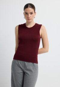 Top sin mangas ajustado burdeos con escote redondo, hecho de una tela suave. Llevado con pantalones a rayas grises. Diseño sencillo.