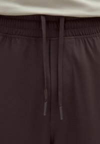 Braune Hose mit elastischem Bund und Kordelzug, glatte Textur und gesteppten Details. Verfügt über eine Seitentasche und verstellbare Bänder.