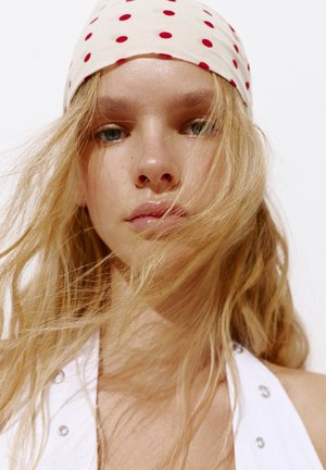Jeune femme aux longs cheveux blonds soufflant sur son visage, portant un haut blanc avec des œillets métalliques et un foulard blanc à pois rouges.