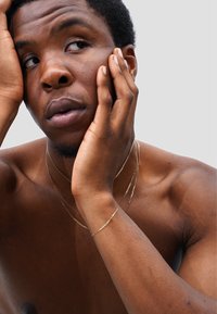 A bare-chested individual with short, dark hair rests their face on their hand, featuring multiple gold chains and a bracelet. The skin tone is medium-dark with visible texture.