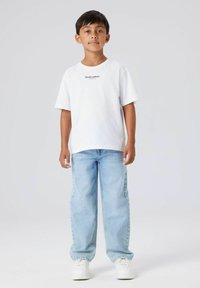 Ragazzo in piedi con jeans azzurri chiari, sneakers bianche e una T-shirt bianca con la scritta "Studio edition" su uno sfondo grigio chiaro semplice.