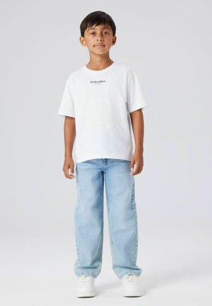 Bernardas stovi šviesiai mėlynuose džinsuose, baltais sportbačiais ir balta marškinėliais, ant kurių yra užrašas "Studio edition", prieš neutralią šviesiai pilką fono.