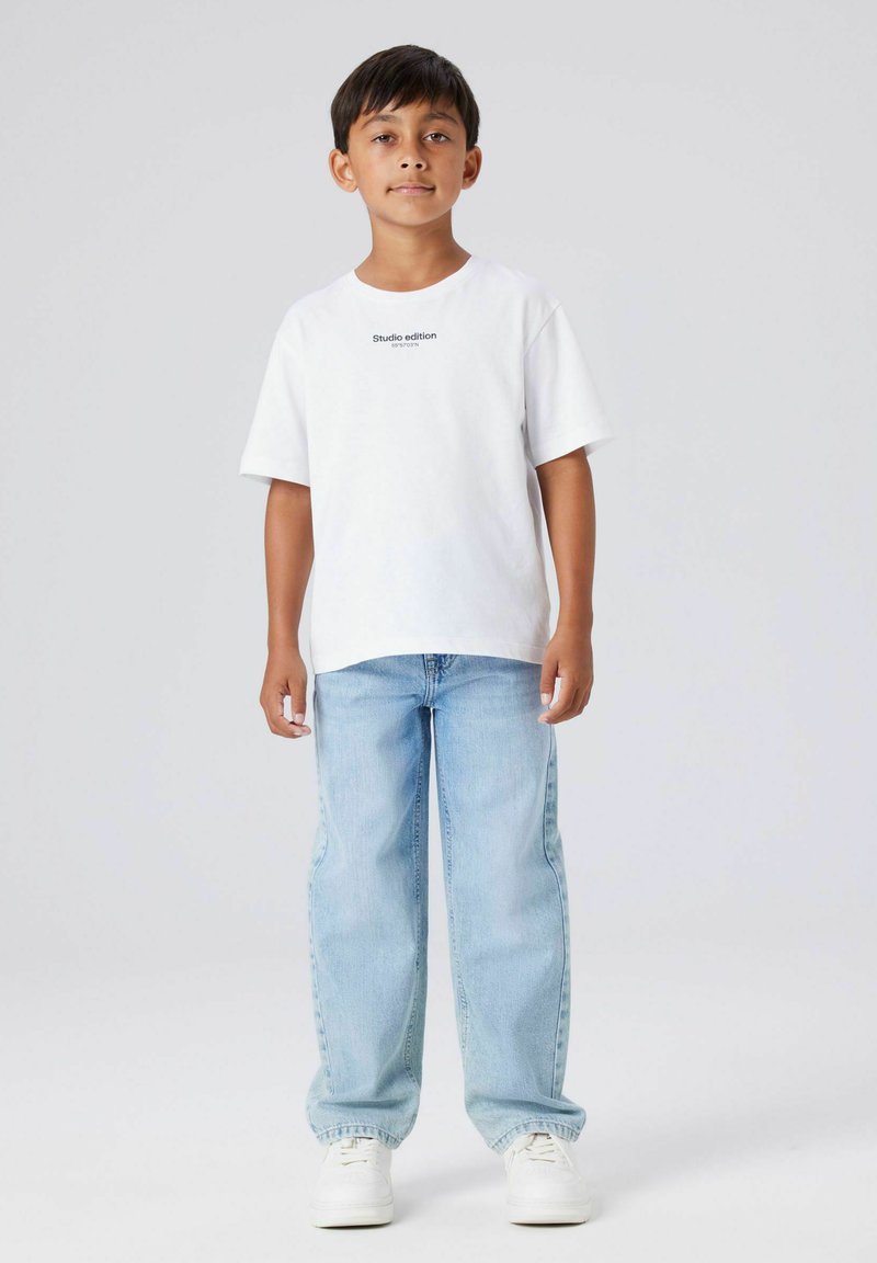 Ragazzo in piedi con jeans azzurri chiari, sneakers bianche e una T-shirt bianca con la scritta "Studio edition" su uno sfondo grigio chiaro semplice.