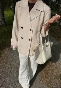 Helebeige kahepoolne jakk mustade nuppudega, kombineeritud valgete laiade pükstega. Kätel tekstuuriga valge käekott.