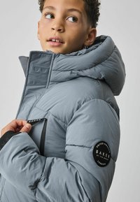 Doudoune grise avec col montant et capuche, dotée d'accents cousus, d'une poche zippée et d'un patch rond de la marque sur la manche.