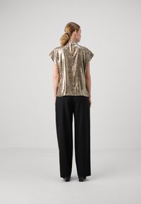 Top in paillettes dorate con collo alto e schiena a chiave, abbinato a pantaloni a gamba larga con righe nere. Texture liscia, finitura scintillante.