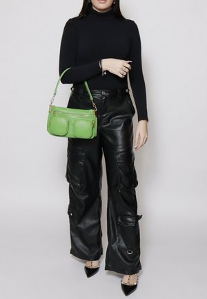 Donna che indossa un dolcevita nero e pantaloni cargo in pelle, che tiene una borsa verde con due tasche frontali e accessori dorati.