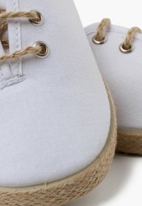 Bílé plátěné boty s juty rope podešvemi, se stříbrnými očky a světle hnědými tkaničkami. Detailní pohled zaměřený na texturu a detaily.