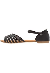 Black sandal with a strappy design, features a flat brown sole, ankle strap with buckle, and a smooth material texture.