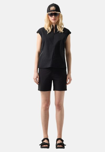 Frau trägt ein schwarzes ärmelloses Oberteil mit Reißverschluss, schwarze Shorts, schwarze Sandalen, eine rückwärts getragene schwarze Kappe und große dunkle Sonnenbrillen, steht vor einem einfarbigen Hintergrund.