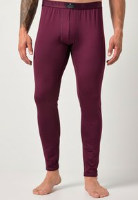 Uomo in leggings termici aderenti color bordeaux, in piedi scalzo contro uno sfondo chiaro e semplice, con un braccio tatuato visibile.