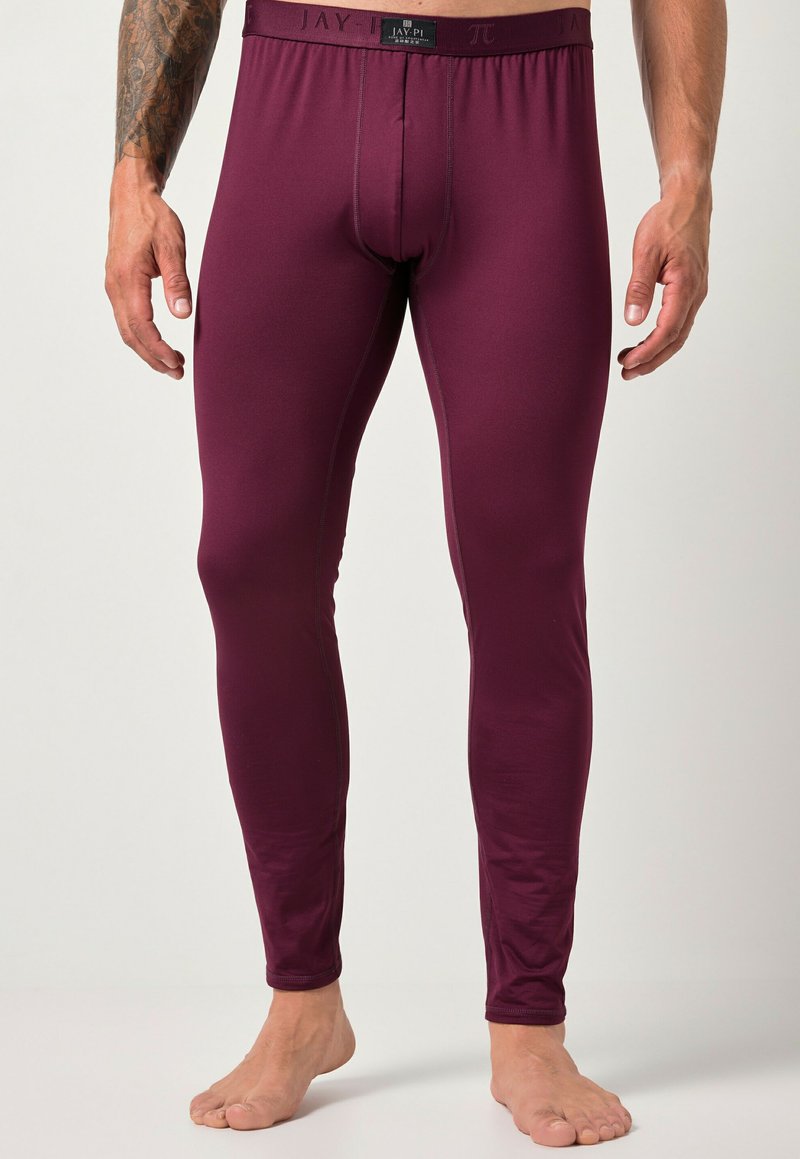 Uomo in leggings termici aderenti color bordeaux, in piedi scalzo contro uno sfondo chiaro e semplice, con un braccio tatuato visibile.