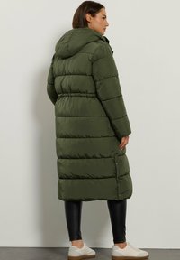 Lang, olivengrøn pufferjakke med hætte, der har et quiltet design, sidelukninger og en afslappet pasform, parret med sorte leggings og lyse sko.