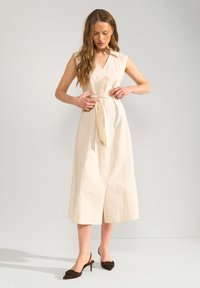Femme ajustant la ceinture d'une robe midi beige sans manches boutonnée, portant des talons pointus marron foncé, debout devant un fond clair uni.