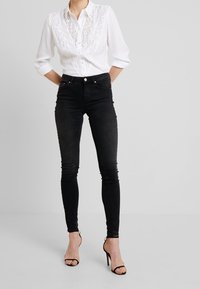 Weißes Button-up-Hemd mit Spitze, kombiniert mit hochtaillierten schwarzen Skinny Jeans und schwarzen Absatzsandalen.