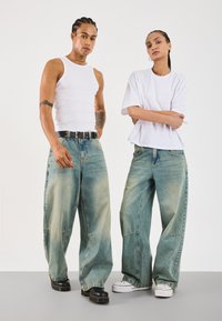 Két személy túlnőtt világoskék farmernadrágot, egy fehér ujjatlan pólót és egy fehér pólót visel, egy szürke felületen állva, egy egyszerű háttér előtt.