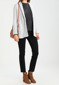 Blazer gris claro de apertura frontal, suéter gris oscuro, pantalones negros de corte slim y botas de tobillo marrones. Modelo sosteniendo un bolso cruzado marrón.