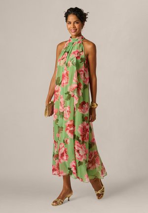Vrouw draagt een mouwloze groene jurk met roze bloemenprint, gouden sandalen, gouden armband en houdt een gouden clutch vast, staand tegen een beige achtergrond.