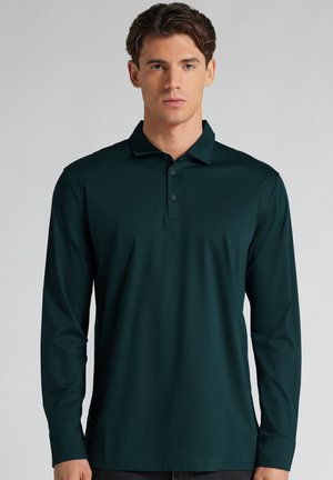 Hombre con cabello corto y castaño que lleva una camiseta polo de manga larga verde oscuro con cuello abotonado, mirando hacia adelante sobre un fondo sencillo.