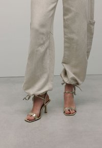 Beige, lichte, taps toelopende broeken met verstelbare manchetten, gecombineerd met metallic gouden hoge hakken met een vierkante neus en dunne banden.
