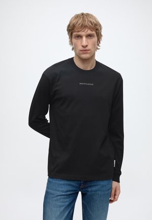 Blondeman die een zwart shirt met lange mouwen draagt met kleine tekst op de borst en blauwe spijkerbroek, staand tegen een effen lichte achtergrond.