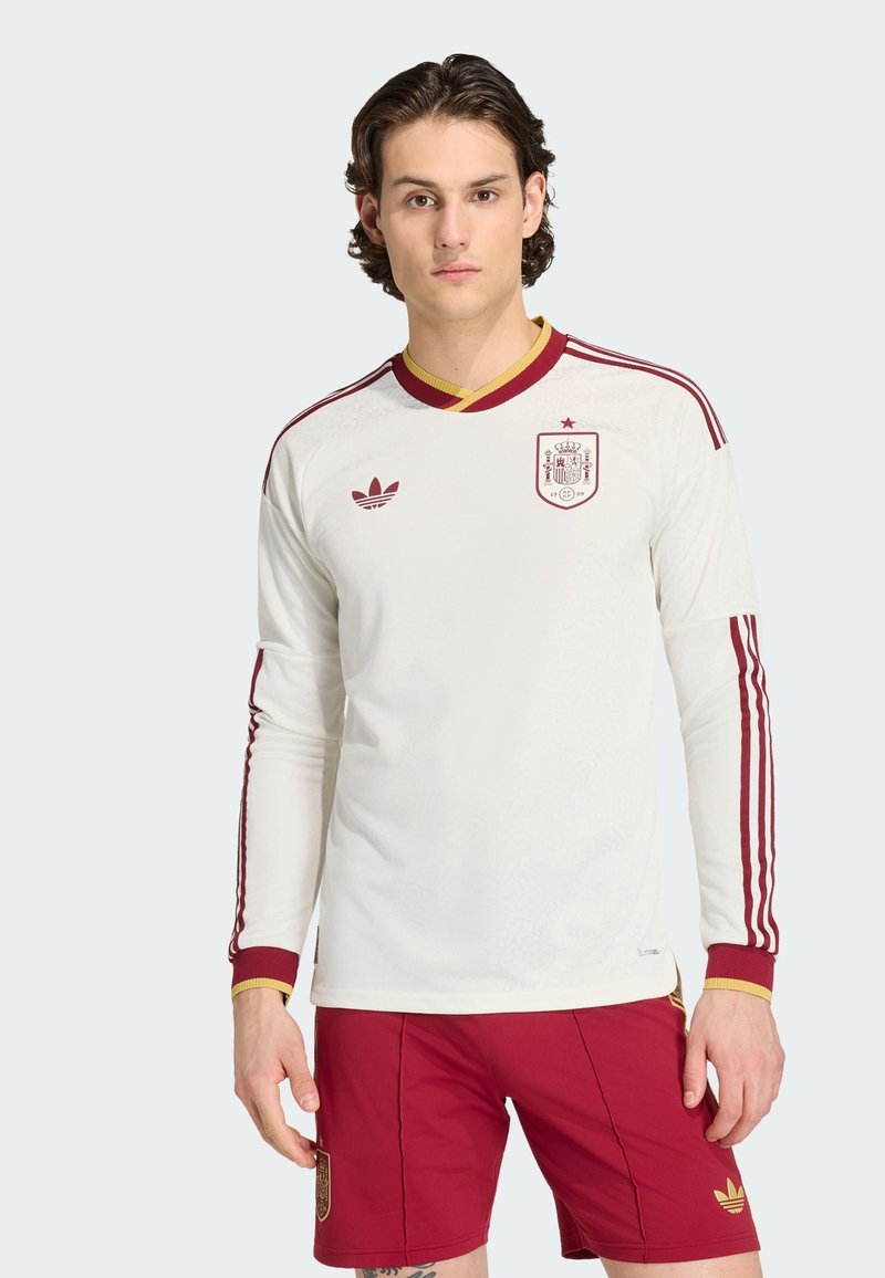 Joven con camiseta blanca de manga larga de fútbol de España con rayas granate y pantalones cortos rojos, de pie contra un fondo claro y liso.