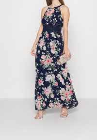 Kvinna i en marinblå halterneck maxiklänning med blommor och rosa rosor, som håller en pärlmeddelad kuvertväska och bär strappy hudfärgade klackar.