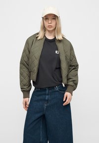 Γυναίκα που φοράει μπεζ ανοιχτό καπέλο μπέιζμπολ, χακί καπιτονέ μπουφάν bomber, μαύρο μπλουζάκι και μπλε τζιν με φαρδιά πόδια σε απλό φόντο.