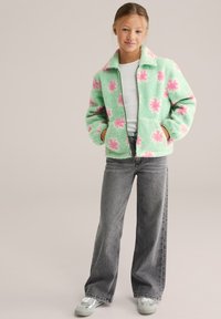 Ragazza giovane in piedi con le mani nelle tasche, indossa una giacca in pile verde menta con motivi floreali rosa, una maglietta bianca, jeans grigi e sneakers.