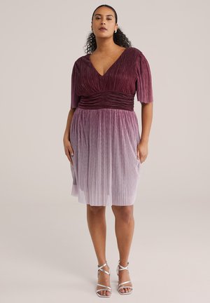 Nő áll, kifinomult bársony redőzött ruhában, amely mélylila felsőrészből világos levendulaszínű szoknyába átmenetet képez, ezüstszínű pántos magassarkúval párosítva.