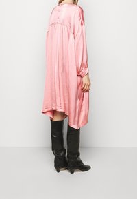 Woman wearing loose, shiny pink dress with long sleeves and three buttons on back yoke, paired with black knee-high heeled boots.