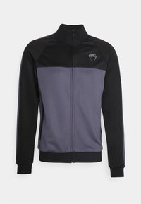 Chaqueta de pista en negro y gris oscuro. Cuenta con una cremallera completa, cuello alzado, paneles de hombro en contraste, bolsillos laterales y un logo en el pecho.