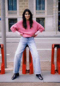 Pull rose moelleux, chemise blanche à manches longues en dessous, jeans larges bleu clair, et bottes noires, assise sur des barrières rouges dans un cadre urbain.