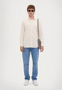 Voľne strihaná béžová košeľa na zapínanie, svetlomodré džínsy, biele tenisky, sivá taška cez rameno a tmavé slnečné okuliare. Minimalistický dizajn, neutrálne farby.