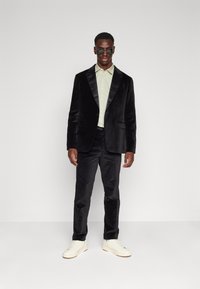 Blazer en velours noir avec des revers en satin, associé à une chemise vert clair, un pantalon noir et des baskets blanches. Comprend des lunettes de soleil rondes.