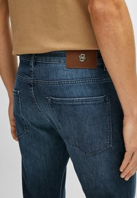 Jean en denim de couleur bleu foncé avec une texture légèrement usée. Présente une patch en cuir marron à l'arrière avec un logo embossé et deux poches arrière.