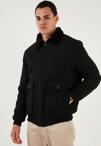 Sort bomberjakke med pelskrave, lynlås foran og to frontlommer med knaplukninger. Ribbet manchetter og talje.