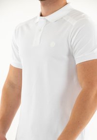 Polo bianco con una texture morbida, caratterizzato da un'apertura a tre bottoni e un piccolo logo sul petto. Maniche corte con polsini a costine.